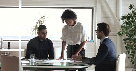 Wall Mural - Focused young african american female employee standing at table, listening to team leader explaining project details. Group of multiracial diverse managers developing corporate strategy in office.