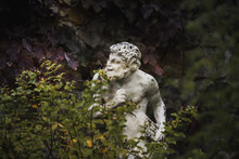 White Statue Sculpture Of Pan The Greek God With A Flute In His Hand Hiding Behind Foliage Looking Cheeky