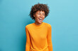 Positive cheerful young African American woman laughs positively and looks aside concentrated happily dressed in casual orange jumper isolated on blue studio background. People emotions concept