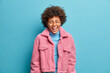 © wayhome.studio - Studio shot of overjoyed dark skinned female model laughs happily and poses with closed eyes dressed in fashionable pink coat expresses sincere emotions isolated on blue background. Clothing concept