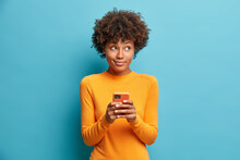 Pensive Delighted Young African American Woman Holds Mobile Phone And Types Message Uses Modern Technologies Dressed In Casual Jumper Isolated On Blue Background. Teenage Girl With Smartphone
