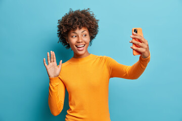 Wall Mural - pretty curly haired positive woman holds modern mobile phone and makes video call waves palm in hell