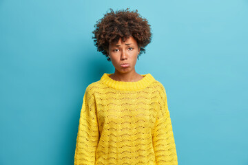 Wall Mural - Attractive offended woman looks with sad frustrated expression purses lower lip and grimaces sadly at camera wears yellow knitted sweater isolated on blue background. Upset dark skinned girl