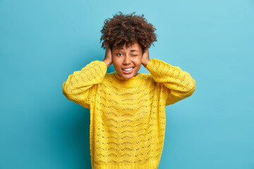 Wall Mural - Horizontal shot of curly haired woman covers ears as hears very loud noise or music winks eye and clenches teeth ignores something noisy dressed in casual yellow jumper poses indoor over blue wall