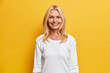 Optimistic glad blonde middle aged woman smiles toothily happy to spend free time in family circle wears casual white jumper isolated on yellow background. Happy senior wrinkled lady indoor.