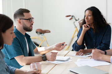 Wall Mural - Confident mentor coach young man wearing glasses training diverse employees at corporate meeting, sitting at table in modern boardroom, colleagues discussing project strategy, results, statistics