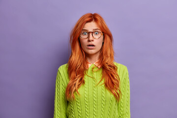 Wall Mural - Young shocked redhead blue eyed woman stares at camera with fearful expression and opened eyes wears green sweater poses against purple background. Ginger female cannot believe in embarrassing news