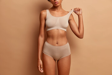 Femine beauty lines concept. Slim woman with dark skin flat stomach wears high waist panties and bra. Female model poses in underwear against brown background