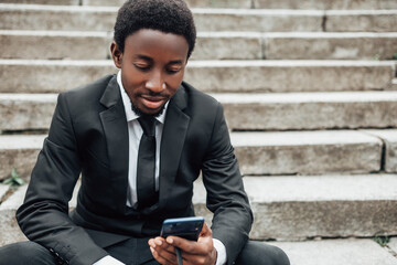 Wall Mural - Young african american businessman wear black suit and sitting on stairs, using his smartphone, surfing the internet.