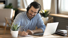 Smiling Young Caucasian Man In Headphones Glasses Sit At Desk Work On Laptop Making Notes. Happy Millennial Male In Earphones Watch Webinar Or Training Course Or Computer, Study Online From Home.