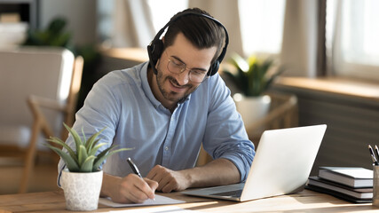 smiling young caucasian man in headphones glasses sit at desk work on laptop making notes. happy mil