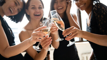 Wall Mural - Close up focus on glasses with champagne or white wine joined together, happy beautiful multiracial female friends in black cocktail dresses celebrating birthday, feeling excited together at party.