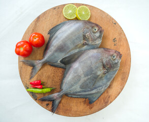 Top view of black pomfret fish dish cooking with various ingredients. Fresh raw fish decorated with lemon slices and tomato slice and curry leaves on a wooden pad, white background.