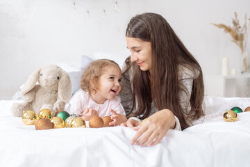 A beautiful curly girl 2 years old with her young mother lie on the bed among garlands and decorations and laugh
