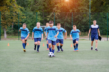 Children running and kicking soccer ball on warming before football training session. Coach in the background watches the training process