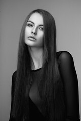 Black and white photo. Frontal portrait of a attractive young woman with long hair and make-up, wear in black bodysuit, looking at camera, over grey background.