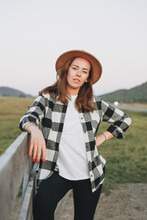 Young Woman In Felt Hat And Plaid Shirt Near Old Wooden Fence On The Mountains Background