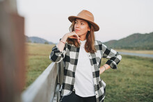 Young Woman In Felt Hat And Plaid Shirt Near Old Wooden Fence On The Mountains Background