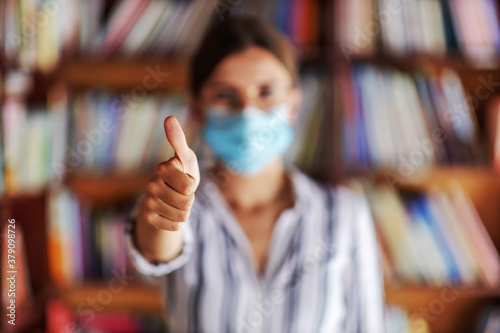 Closeup of college girl standing in library and holding thumbs up. She is having face mask to prevent corona virus from spreading.