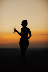 Wall Mural - Silhouette of slim girl with wine glass on colorful sunset. Copy space.