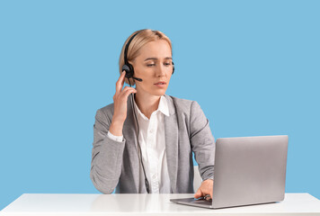 Attractive female helpline operator with headset and laptop answering client's call at table against blue background