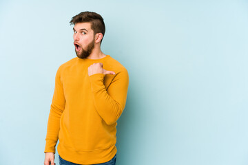 Wall Mural - Young caucasian man isolated on blue background points with thumb finger away, laughing and carefree.