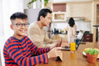© DragonImages - Smiling Asian preteen boy in glasses studying online at kitchen table, his father working on laptop nearby