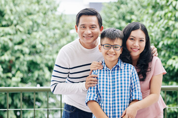 Poster - Portrait of joyful Asian mother, father and their preteen kid smiling and looking at camera when standing on apartmernt balcony