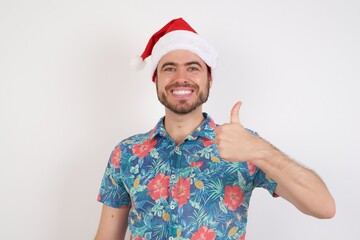 Young caucasian man wearing hawaiian shirt and Santa hat over isolated white background doing happy thumbs up gesture with hand. Approving expression looking at the camera showing success.