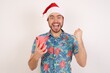© Roquillo - Positive Young caucasian man wearing hawaiian shirt and Santa hat over isolated white background holds modern cell phone connected to headphones, clenches fist from good emotions, exclaims with joy,