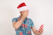 © Roquillo - Young caucasian man wearing hawaiian shirt and Santa hat over isolated white background looking at smart phone feeling sad holding hand on face.