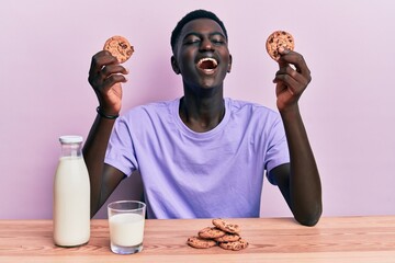 Young african american man drinking a glass of fresh milk with cookies angry and mad screaming frustrated and furious, shouting with anger looking up.