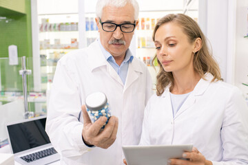 Poster - Senior drugstore worker in whitecoat and eyeglasses showing pills to assistant