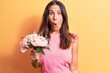 © Krakenimages.com - Young beautiful brunette woman holding bouquet of pink flowers over yellow background scared and amazed with open mouth for surprise, disbelief face