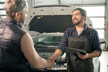 Happy young technician of car service shaking hand of male client after repair