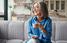 Happy Beautiful Relaxed Mature Older Adult Grey-haired Woman Drinking Coffee Relaxing On Sofa At Home. Smiling Stylish Middle Aged 60s Lady Enjoying Resting Sitting On Couch In Modern Living Room.