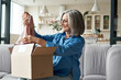 © insta_photos - Happy middle aged woman buyer opening parcel box at home. Smiling old mature female customer shopper receiving online shop purchase buying clothes unpacking delivery postal shipping package on couch.