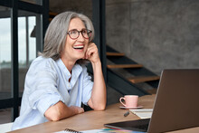 Happy Stylish Mature Middle Aged Business Woman Wearing Glasses Laughing Sitting At Workplace Desk With Laptop. Cheerful Positive Older Grey-haired Businesswoman Having Fun Working Alone In Office.