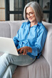 © insta_photos - Happy 60s older mature middle aged adult woman holding laptop using computer sitting on couch at home. Smiling elegant senior grey-haired lady spending time with technology device in living room.