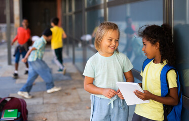 Two cute preteen schoolgirls talking friendly near school building during break between lessons on warm autumn day