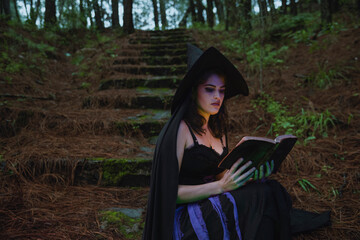 Halloween witch young girl wicca wearing black costume witches holding a old book for a spells in a dark forest  with magic lights