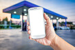 Hand holding a white smartphone with mockup blank screen blur background of twilight gas station at sunset. Touch screen mobile phone at the petrol station.