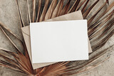 Fototapeta Boho - Summer stationery still life. Closeup of blank card mock-up and craft envelope on dry palm leaf. Grunge beige concrete background. Flat lay, top view. Tropical vacation concept. Moody boho design.