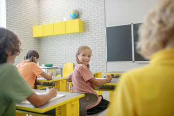 Cute girl sitting at the desk in the classroom and smiling to her classmate