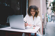 © BullRun - Focused young woman taking notes in front of laptop in summer area of cafeteria