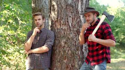 Wall Mural - Two Strong men lumberjacks with an ax in a plaid shirt. Handsome lumberjacks with a large ax examines the tree before felling. Firewood as a renewable energy source.