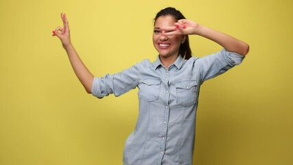 Wall Mural - young woman in blue denim shirt making dance gestures, pointing fingers, holding fists in the air, dancing and celebrating on yellow background