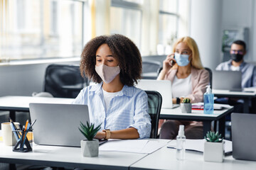 Social distancing at coronavirus outbreak situation. Pretty african american lady in protective mask typing on laptop, working for company support center