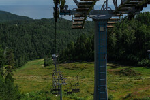Cable Car Construction With Empty Seats Among Green Trees, Grass Field In Forest On Hill, Sunny Summer Blue Sky With White Clounds, Sea On Horizon