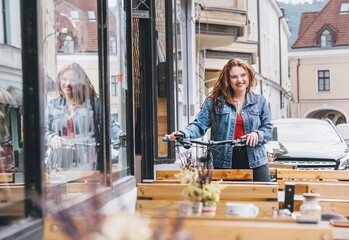 Wall Mural - Portrait of a smiling red curled long hair caucasian woman on the restaurant terrace on the city street walking with a bicycle. Natural people beauty concept image.